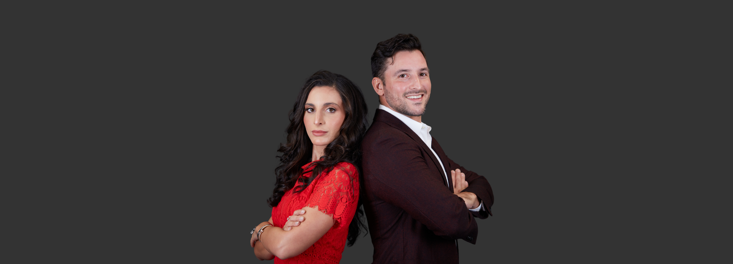 Peter Sakaris, founder of Sakaris Jewelers, and Daria Sakaris, his wife and business partner, standing back-to-back against a dark gray background. Peter, wearing a maroon blazer and white shirt, smiles confidently, while Daria, in a red lace dress, gazes directly at the camera with a poised and professional expression. Together, they represent the leadership and dedication behind Sakaris Jewelers.